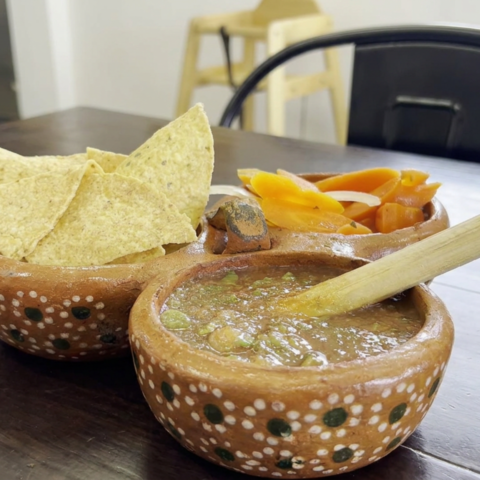 Traditional Mexican clay bowls hold tortilla chips, chunky green salsa, and orange-red salsa on a wooden restaurant table.
