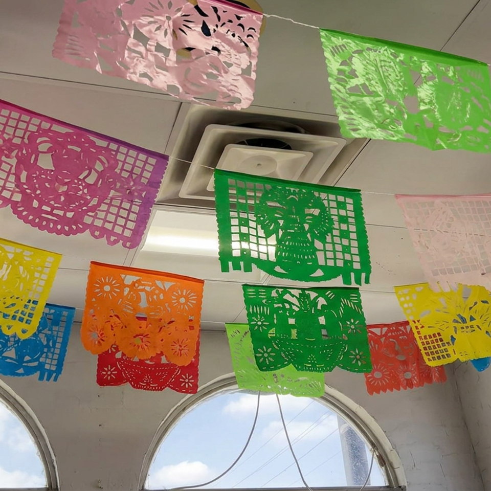 Colorful papel picado banners hang across a restaurant ceiling above arched windows and bright daylight.